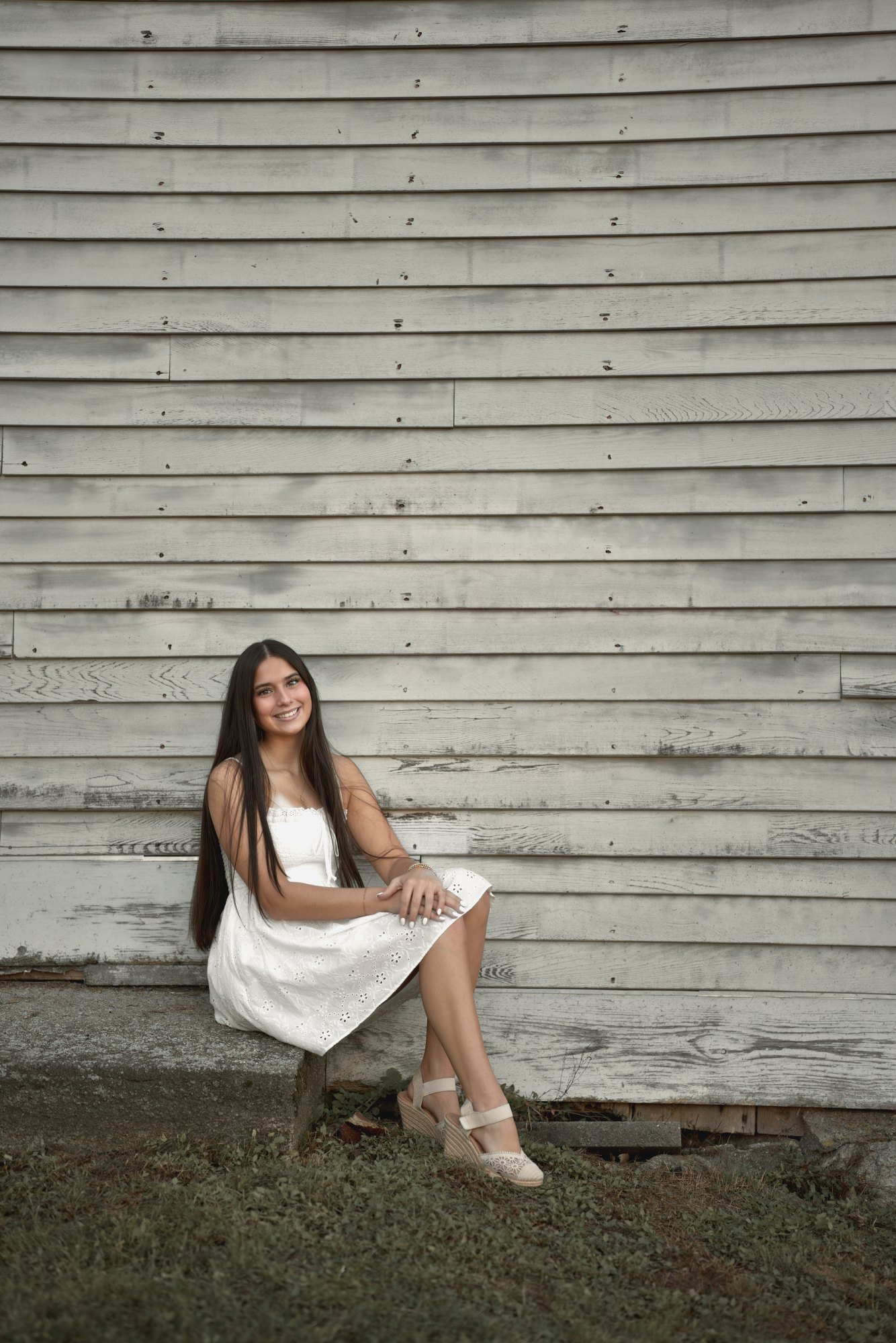 Young woman in a white eyelet dress sitting against weathered grey clapboard