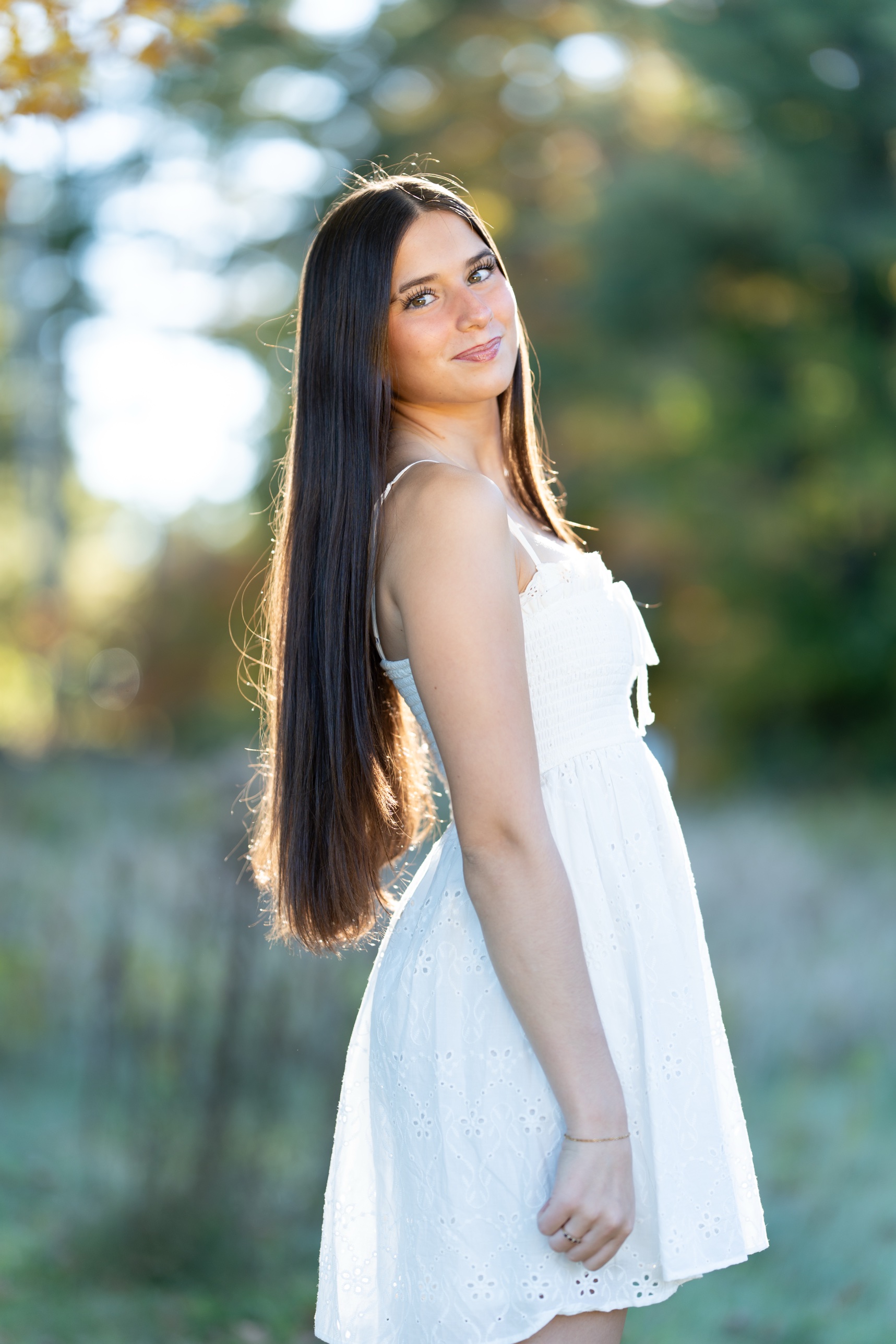 Young woman with long dark hair in a white eyelet sundress, glancing back over her shoulder in soft backlight